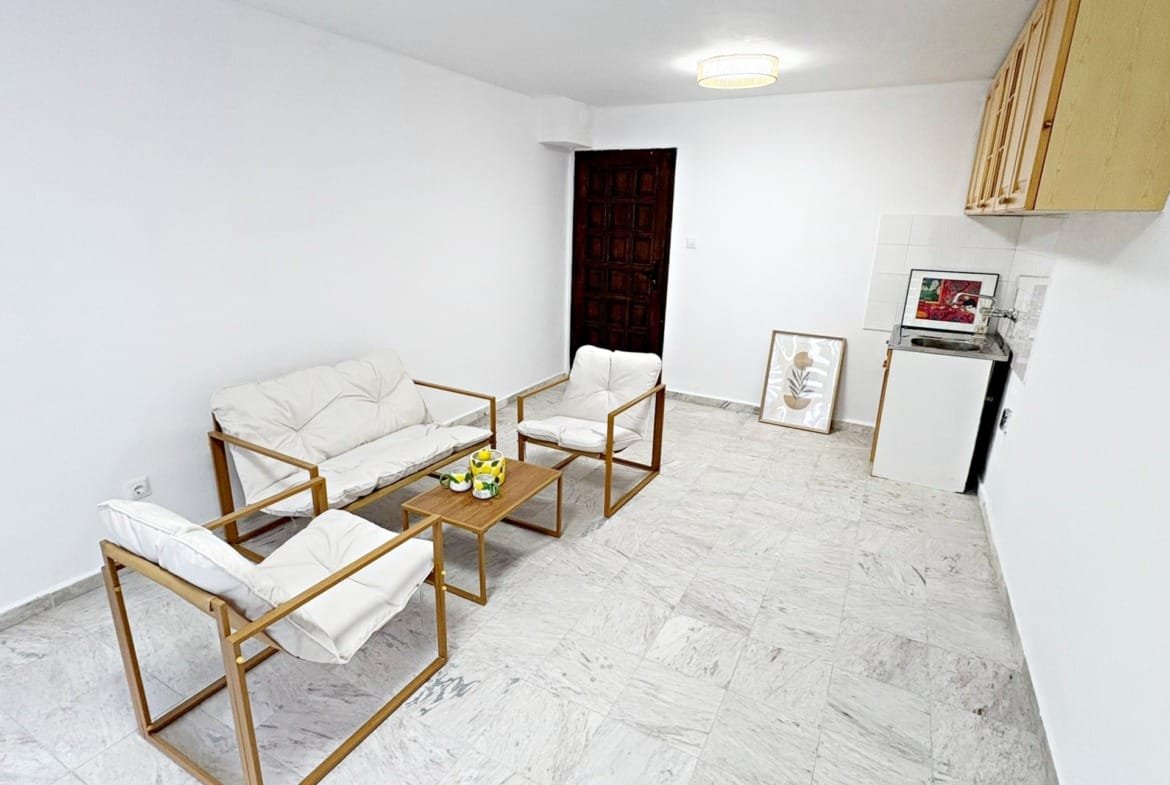 Современная квартира в Болгарии с белыми стенами, мягкой мебелью и кухонным уголком, подходит для покупки или аренды.