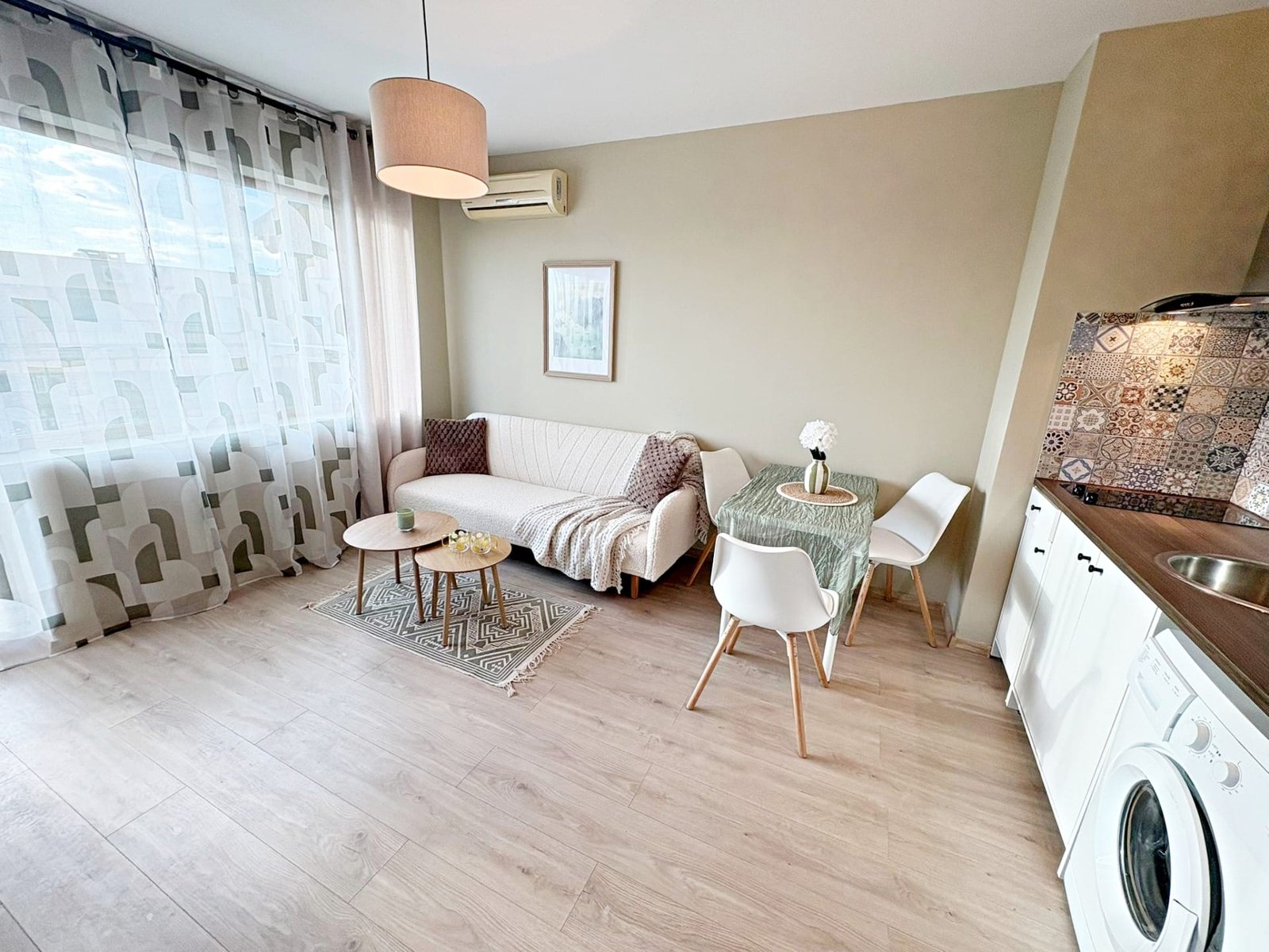 Современный интерьер квартиры в Болгарии с уютной гостиной, столом и кухонным оборудованием. Светлые тона и удобная мебель.