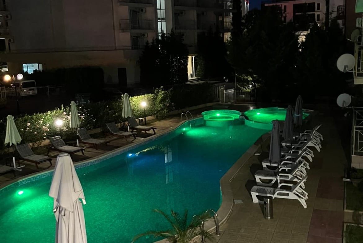 Современный бассейн с подсветкой, окруженный шезлонгами и зонтиками, в жилом комплексе Болгарии. Ночная атмосфера курорта.
