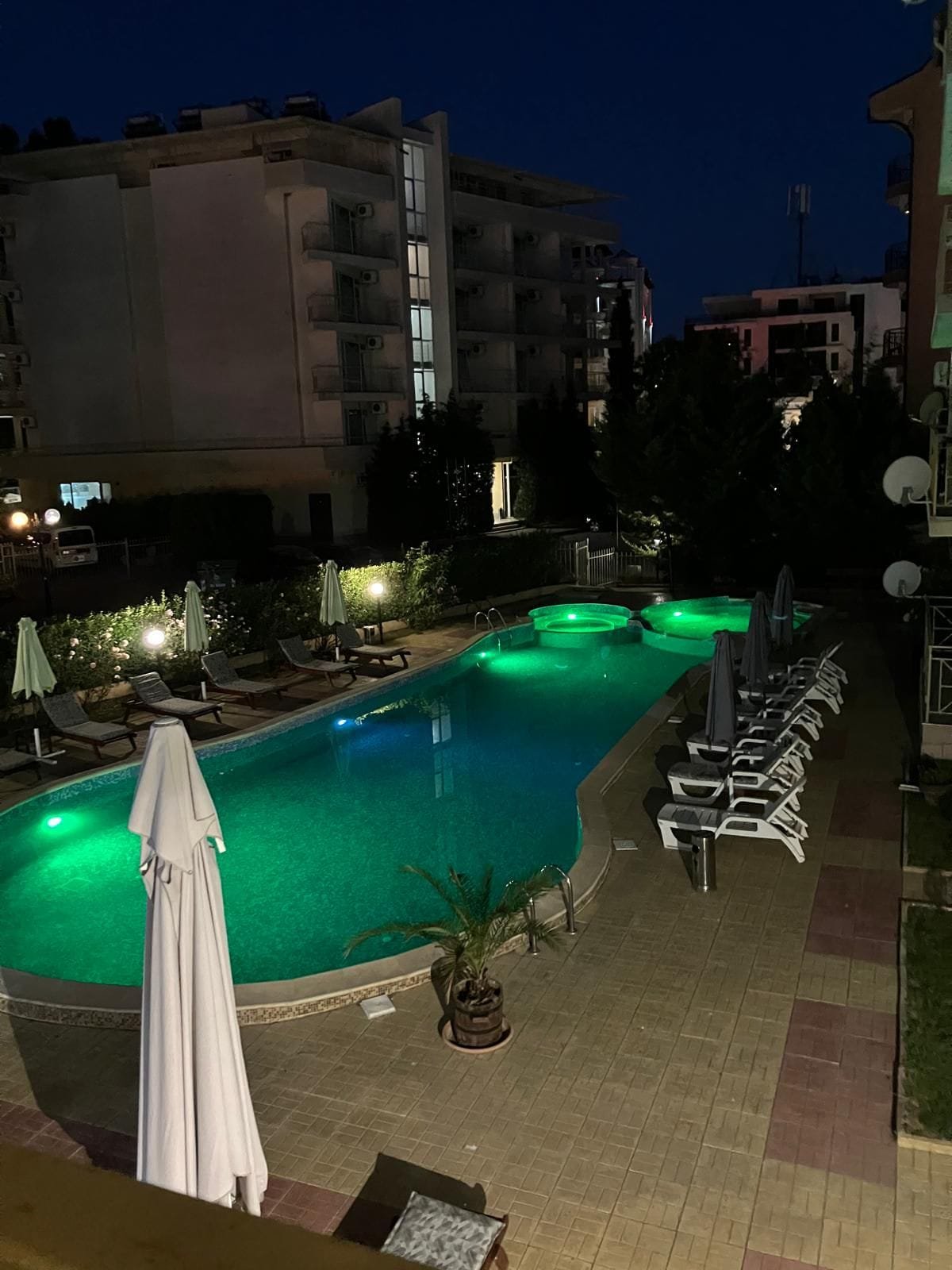 Уютный отель с бассейном ночью и зелеными огнями. Современный бассейн с подсветкой, окруженный шезлонгами и зонтиками, в жилом комплексе Болгарии. Ночная атмосфера курорта.