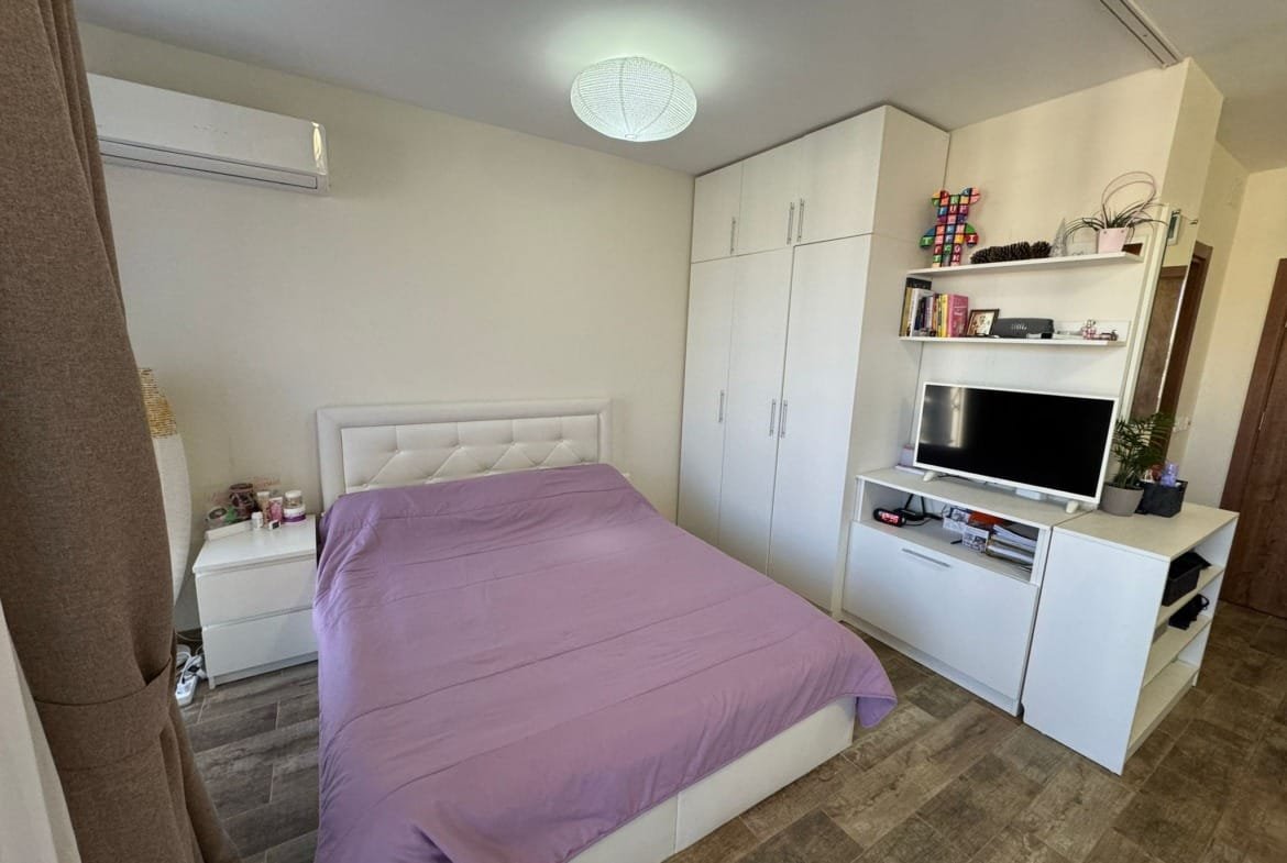 Современная спальня с белой мебелью, кроватью с фиолетовым покрывалом и телевизором на полке, идеальная для жизни в Болгарии.