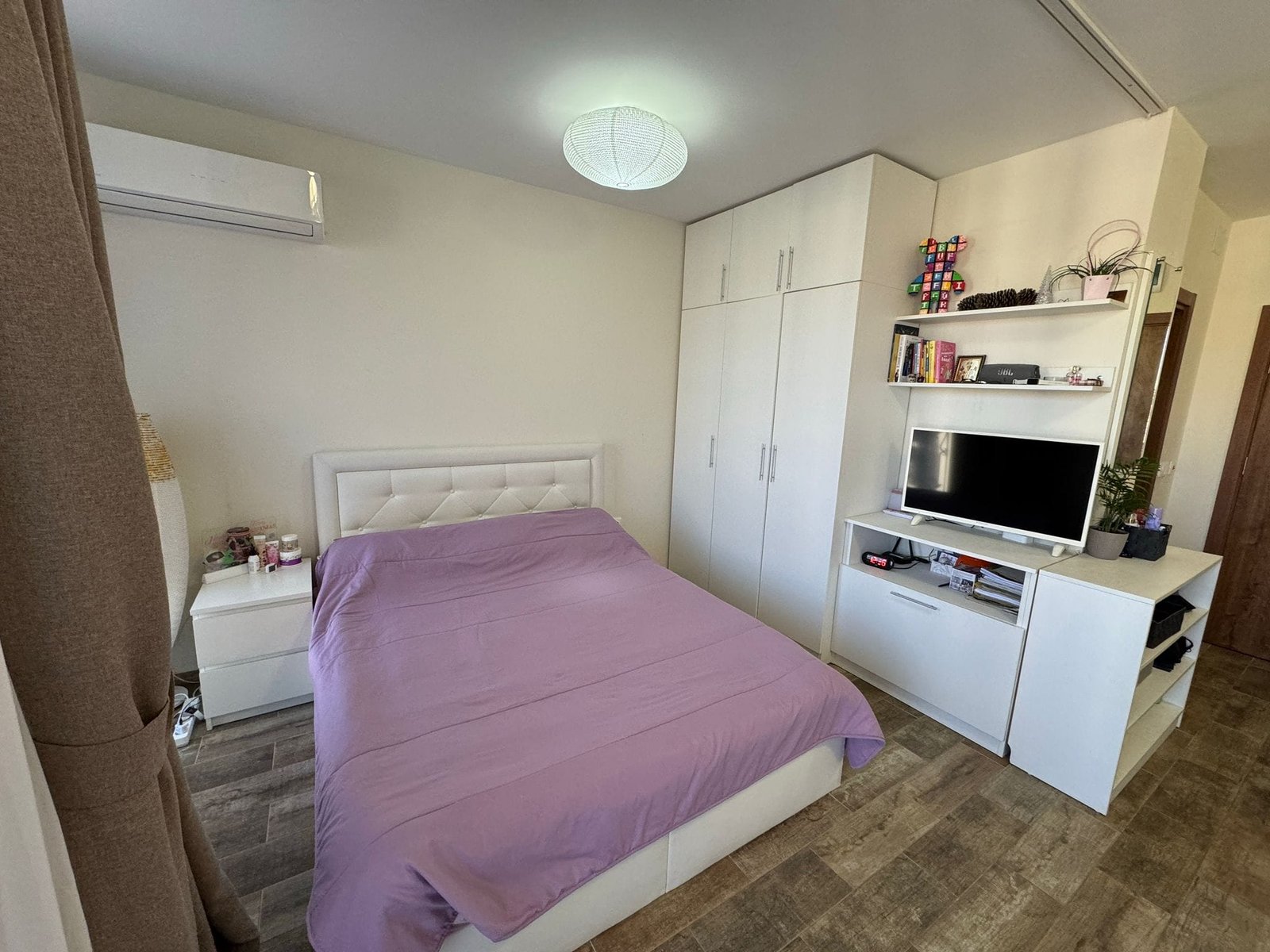 Современная спальня с белой мебелью и фиолетовым покрывалом Современная спальня с белой мебелью, кроватью с фиолетовым покрывалом и телевизором на полке, идеальная для жизни в Болгарии.