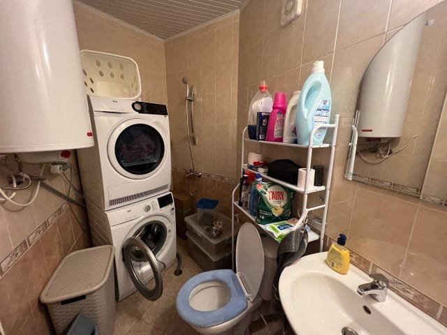 Стиральная машинка, сушилка и унитаз в небольшой ванной с плиткой, полками с чистящими средствами и раковиной.