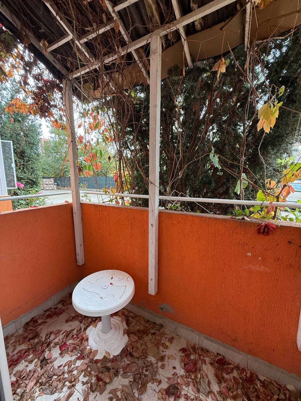 Терраса с белым столом и оранжевыми стенами, окружённая зеленью и осенними листьями. Спокойная обстановка для отдыха.