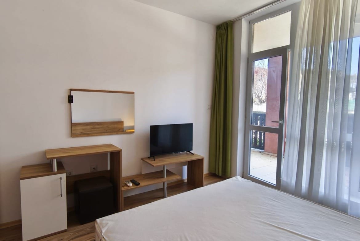 Современная квартира в Болгарии с уютной спальней, телевизором и балконными дверями, оформленная в светлых тонах.