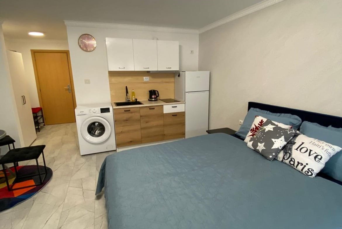 Современная студия с кухонной зоной, стиральной машиной и уютной кроватью, идеальная для покупки или аренды в Болгарии.