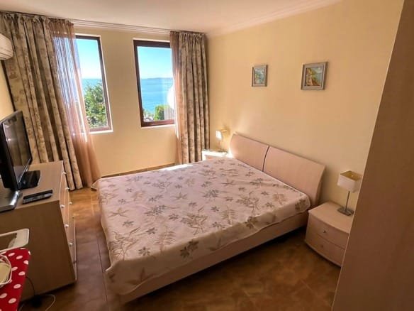 Современная спальня с двуспальной кроватью и видом на море, стены светлых тонов, уютная атмосфера для отдыха в Болгарии.