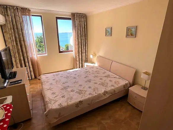 Современная спальня с двуспальной кроватью и видом на море, стены светлых тонов, уютная атмосфера для отдыха в Болгарии.