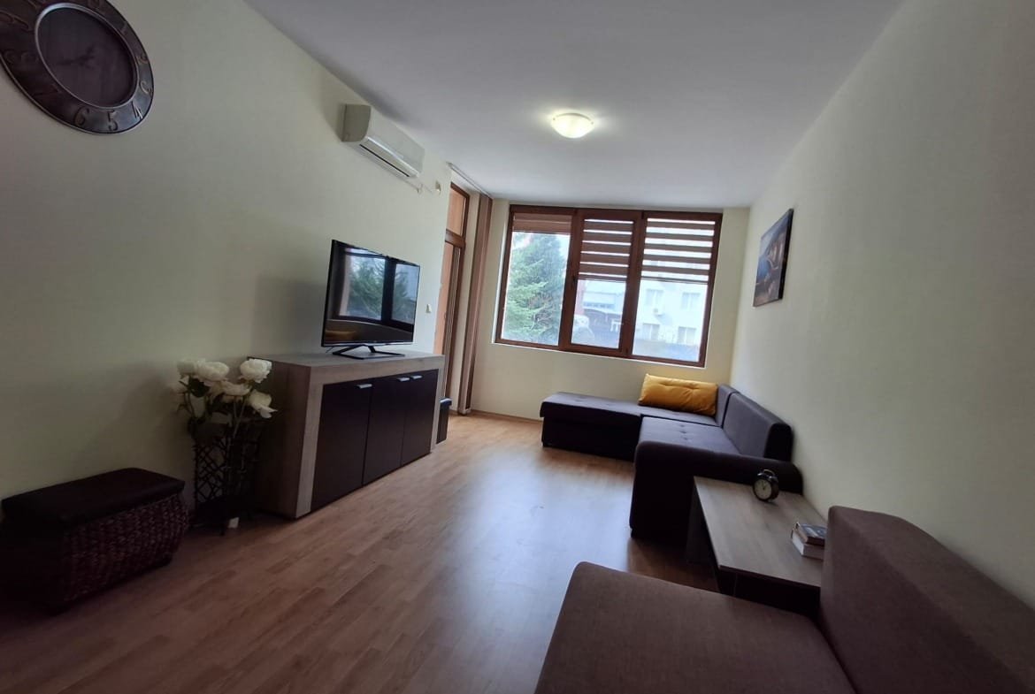 Современная гостиная с телевизором, диваном, столом и кондиционером, светлые тона, большое окно с жалюзи.