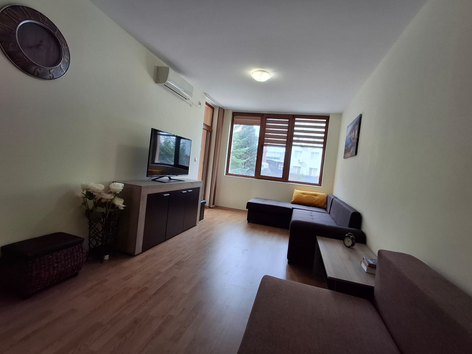 Современная гостиная с телевизором, диваном, столом и кондиционером, светлые тона, большое окно с жалюзи.