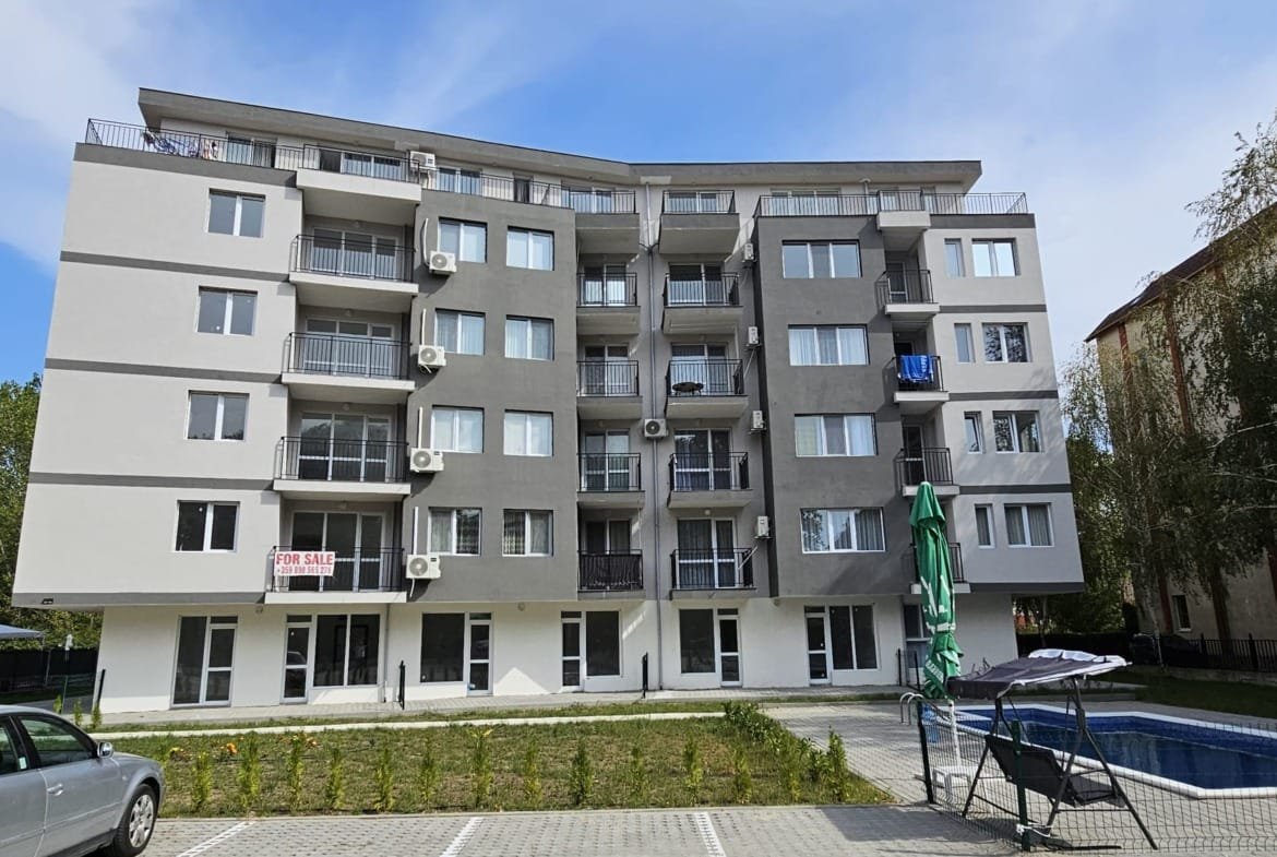 Современный многоквартирный дом в Болгарии с бассейном, парковкой и объявлениями о продаже недвижимости.