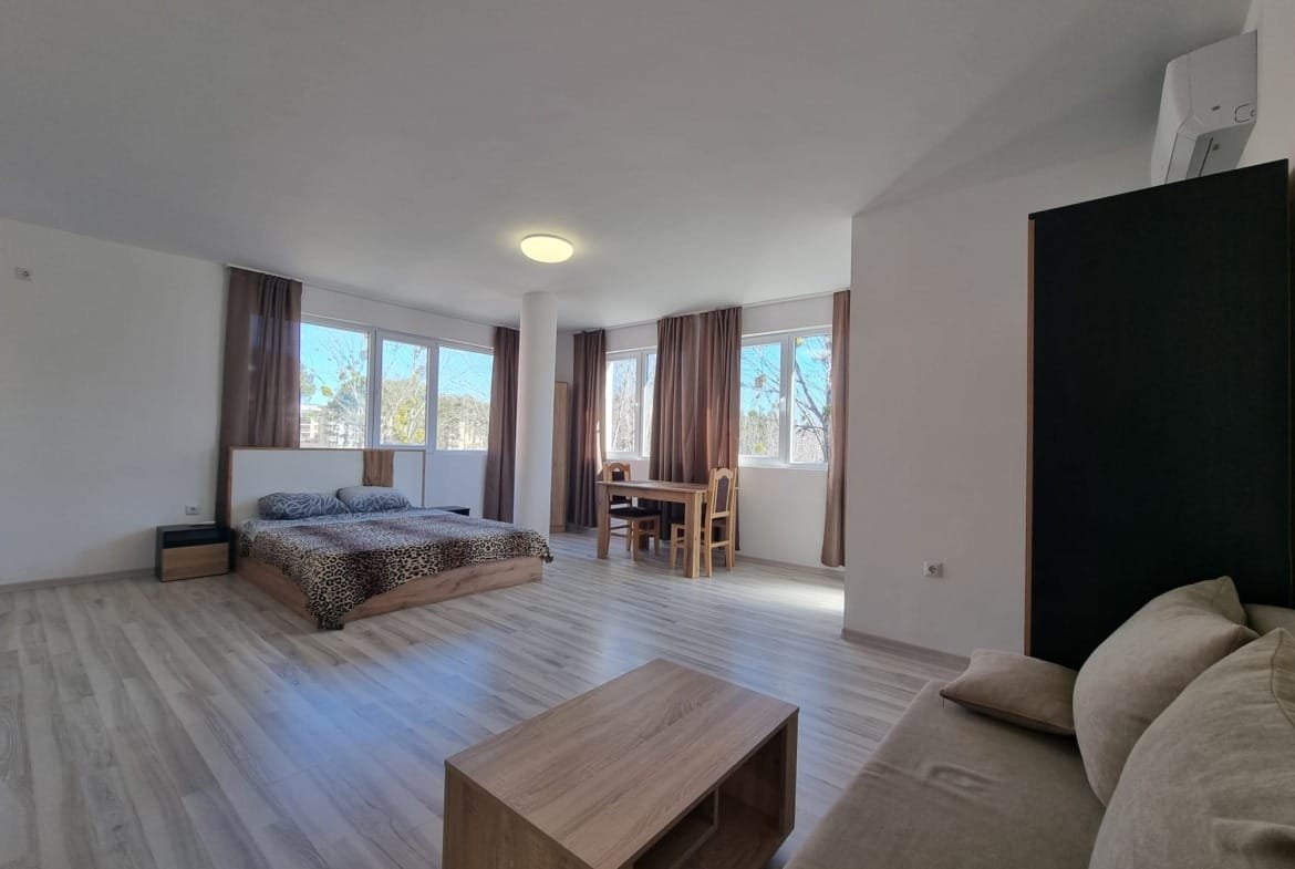 Современный интерьер квартиры в Болгарии с двумя окнами, кроватью, обеденным столом и зоной отдыха. Просторное помещение с естественным светом.