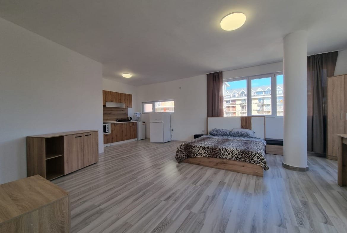 Современная студия в Болгарии с кухней и бытовой техникой, светлым интерьером и большим окном. Подходит для комфортного проживания.