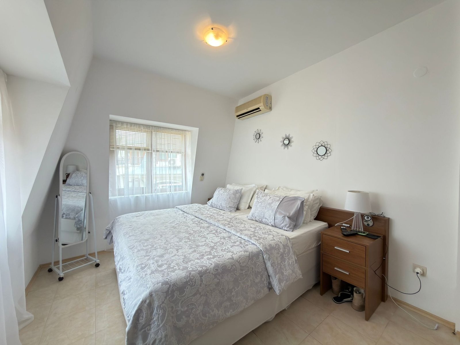 Светлая спальня с двуспальной кроватью, комодом и окном, оформленная в современном стиле, подходит для отдыха в Болгарии.