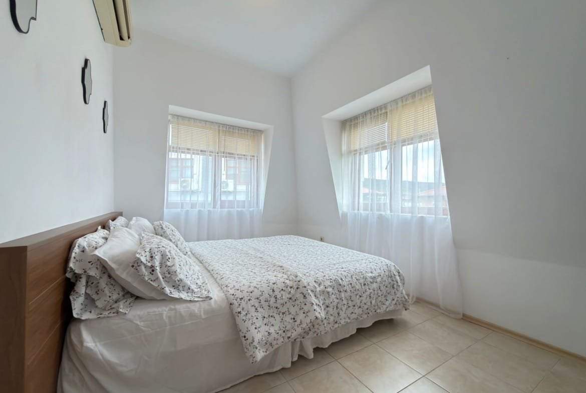 Современная спальня с светлыми стенами, большой кроватью и тройными окнами, идеально подходящая для покупки недвижимости в Болгарии.