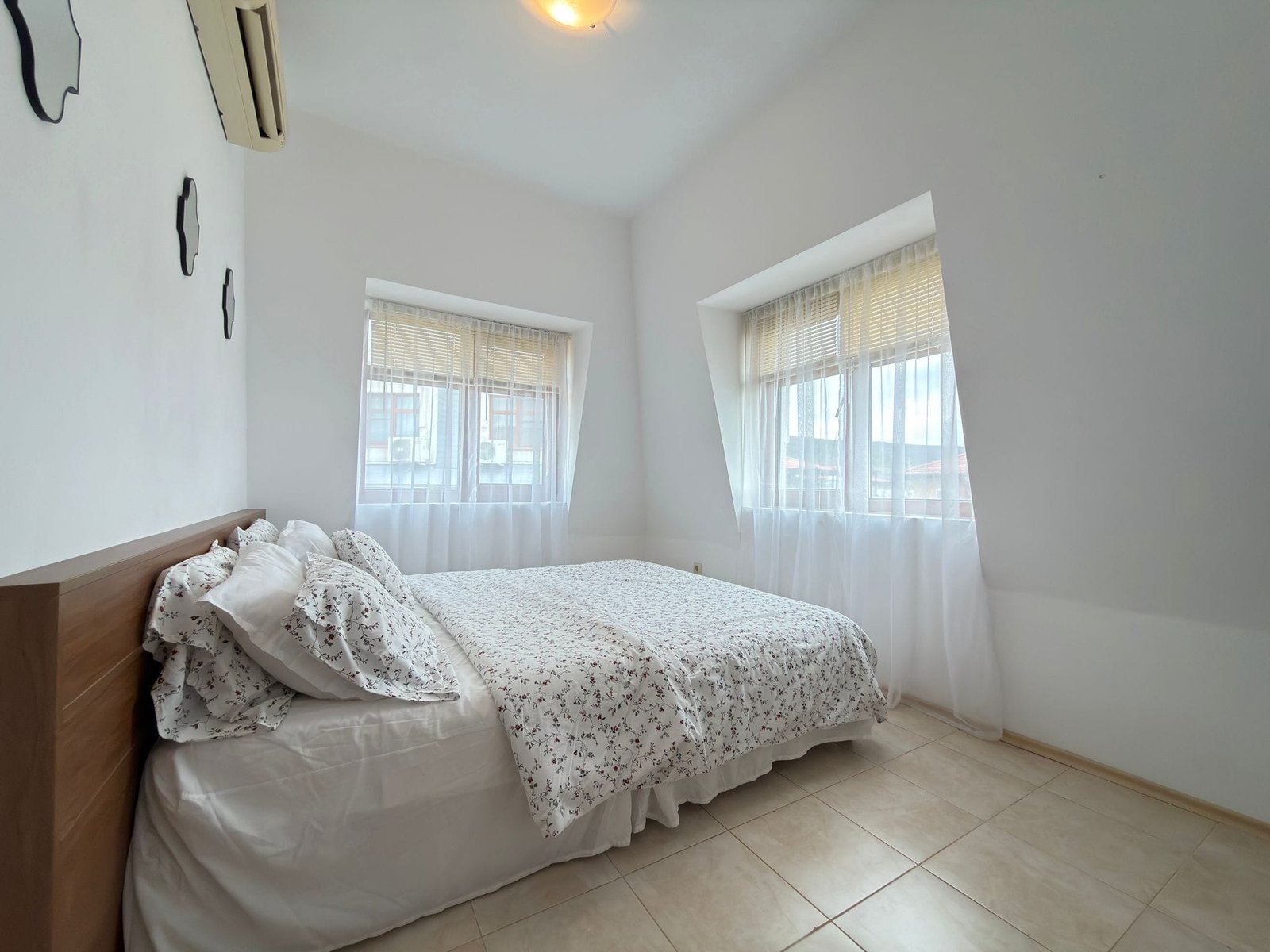 Современная спальня с светлыми стенами, большой кроватью и тройными окнами, идеально подходящая для покупки недвижимости в Болгарии.