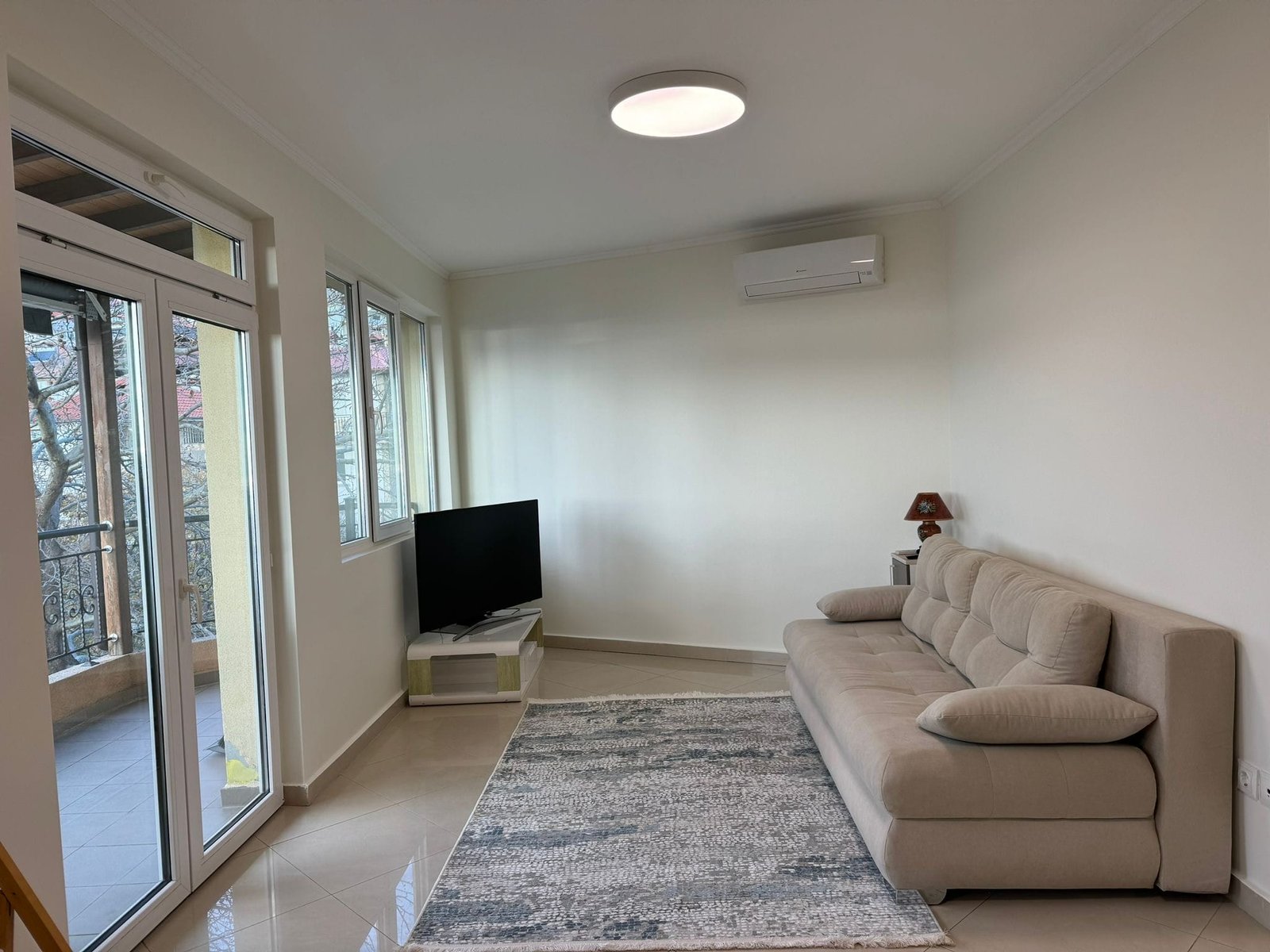 Современная гостиная с диваном и телевизором в светлом интерьере Современная гостиная с диваном, телевизором и светлым интерьером, идеальная для покупки недвижимости в Болгарии.