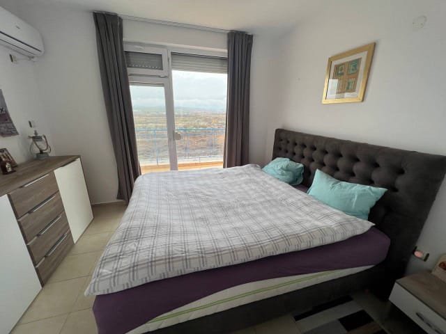 Современная спальня с двуспальной кроватью, окнами с видом и светлыми постельными принадлежностями. Идеально для покупки недвижимости в Болгарии.