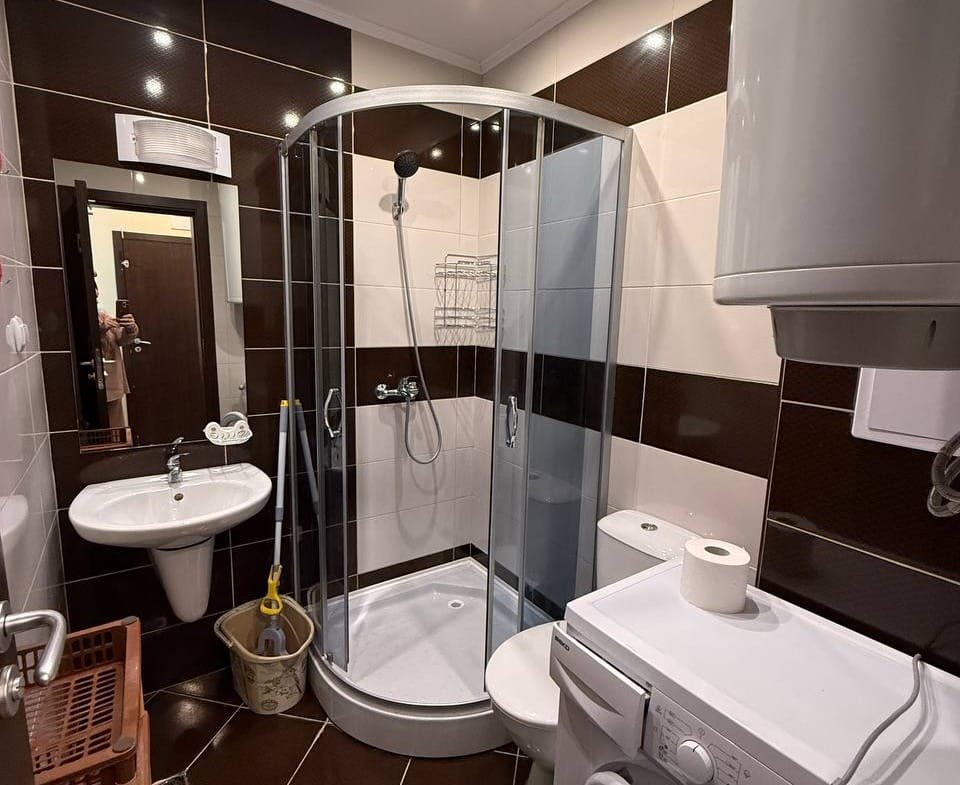 Современная ванная комната с душевой кабинкой, умывальником, стиральной машиной и гладкими темными плитками.