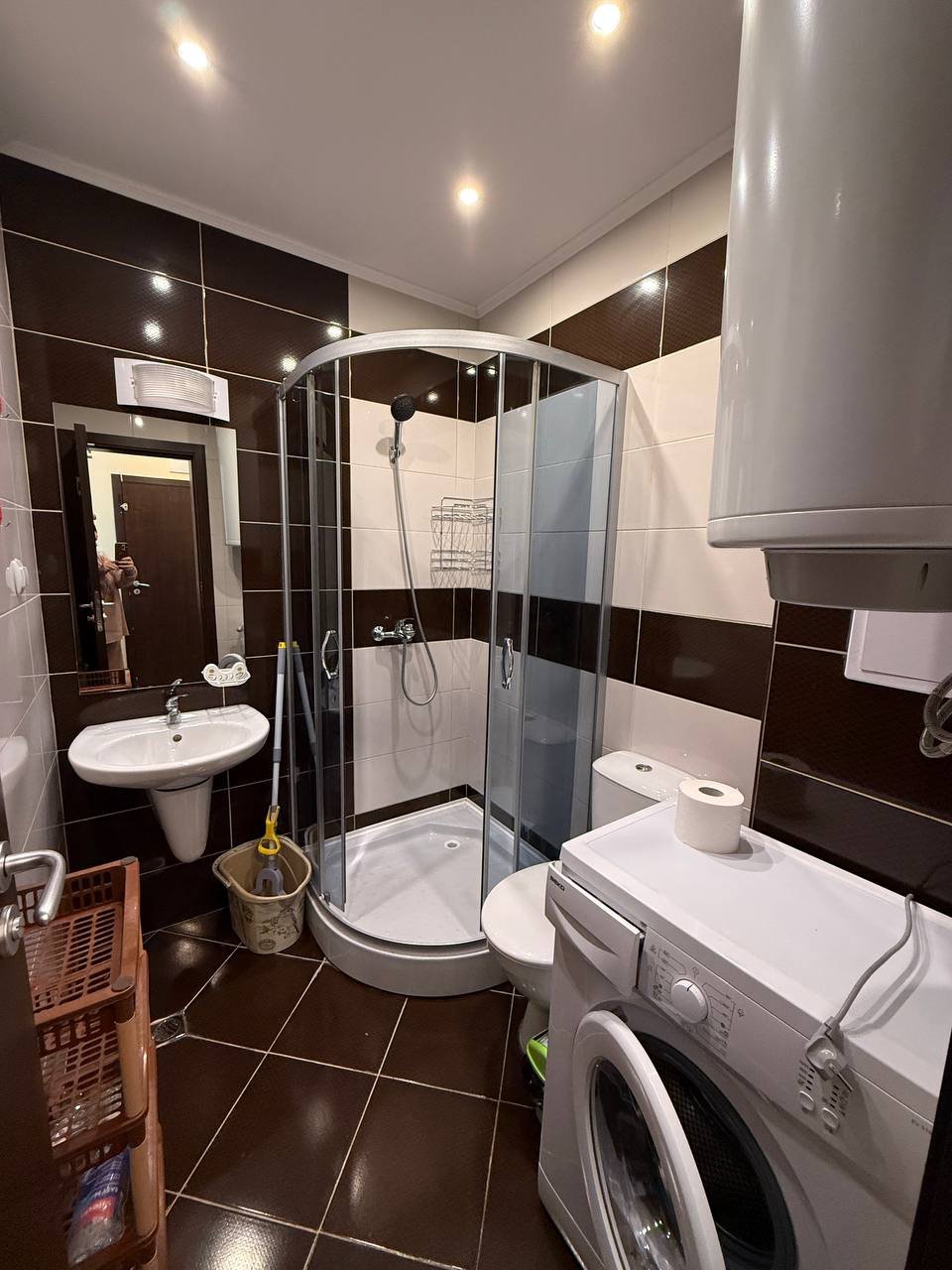 Современная ванная комната с душевой кабинкой, умывальником, стиральной машиной и гладкими темными плитками.