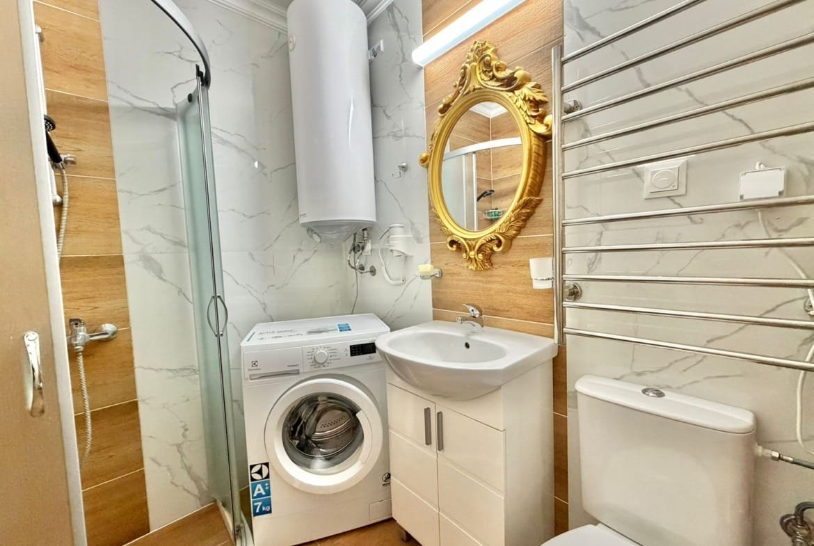 Санузел в квартире с душевой кабиной, стиральной машиной и золотым зеркалом на стене, стильный и современный.