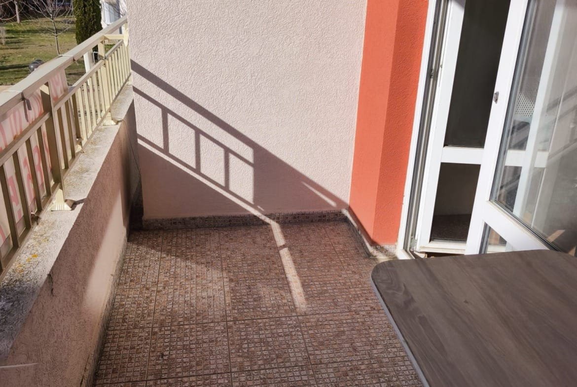 Балкон с плиточным полом и деревянным столом, солнечный свет, вид на зелёный двор и соседние здания в Болгарии.