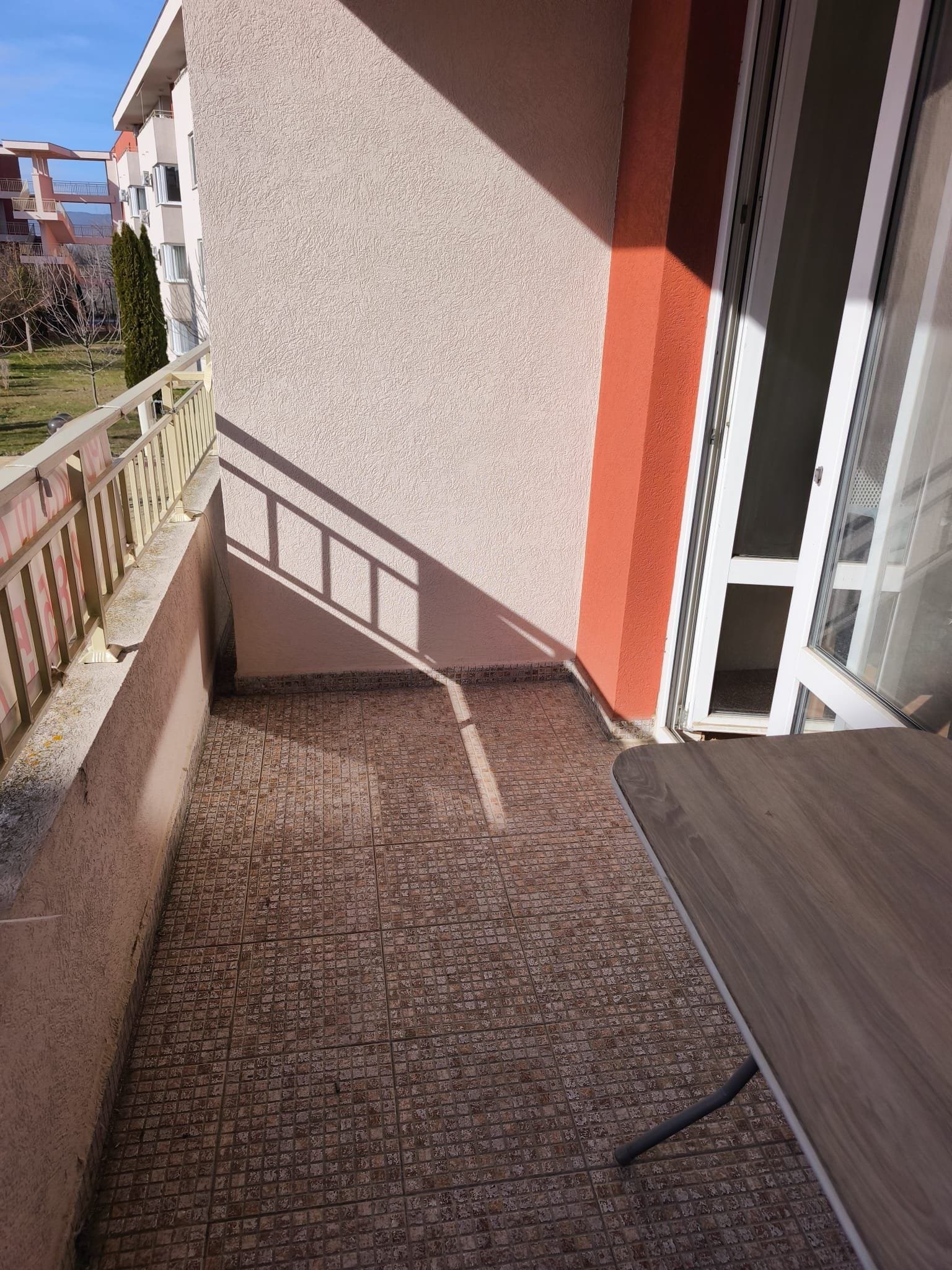 Балкон с солнечными тенями и столом на улице. Балкон с плиточным полом и деревянным столом, солнечный свет, вид на зелёный двор и соседние здания в Болгарии.