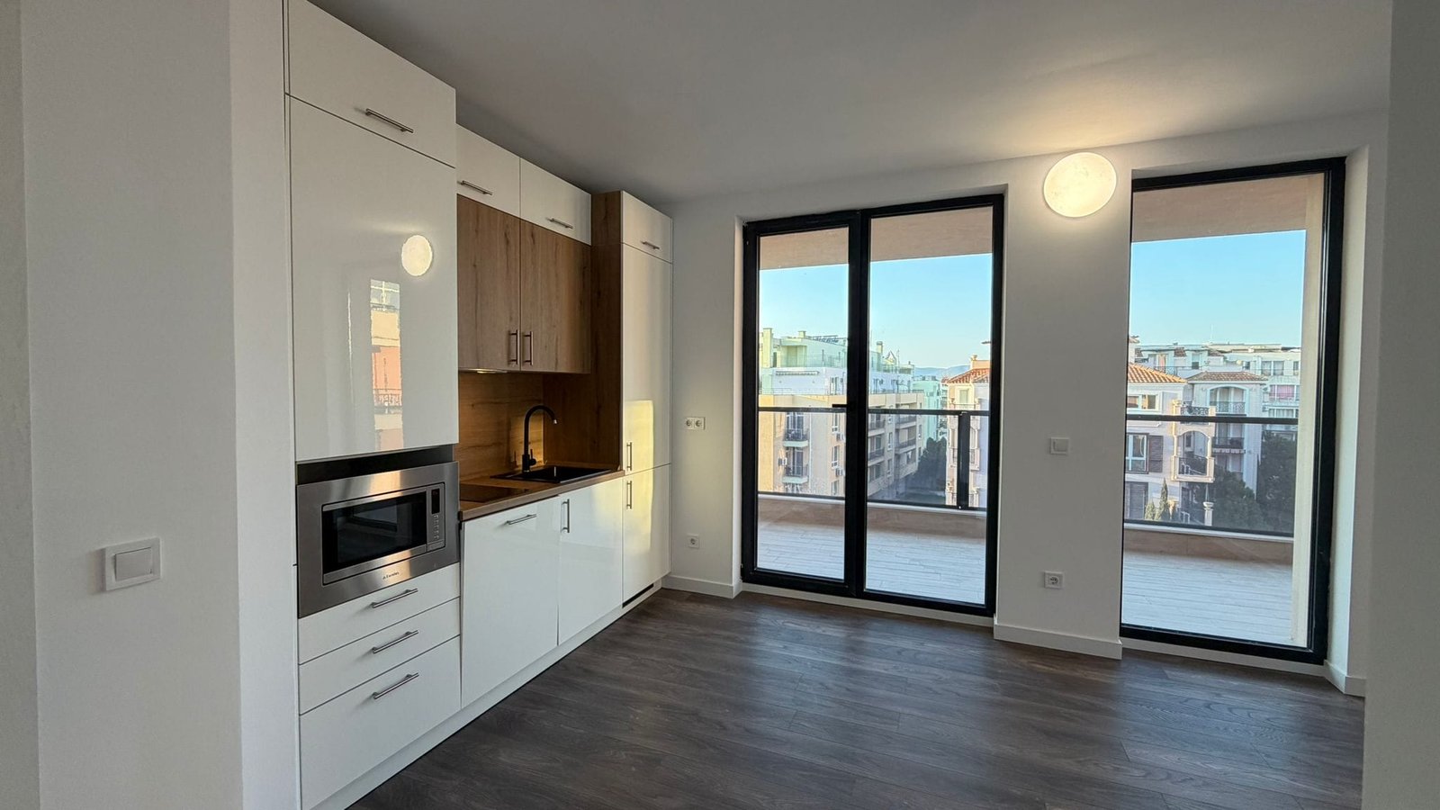 Современная кухня в новой квартире с балконом и солнечным светом. Современная кухня с белыми шкафами и деревянными акцентами, большие окна с видом на городской пейзаж в Болгарии.