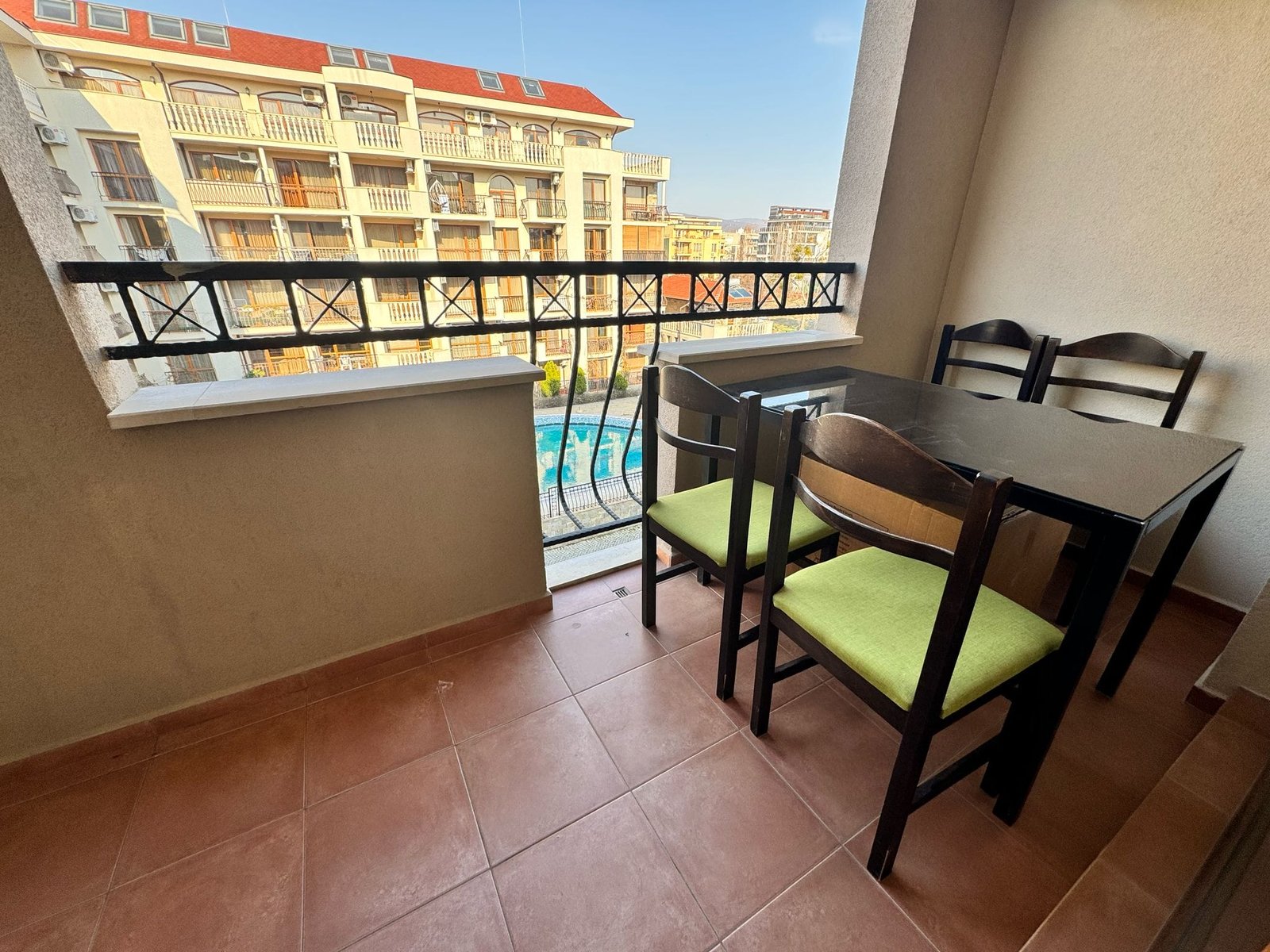 Балкон с набором мебели: стол и четыре стула, вид на плавательный бассейн и жилые комплексы в Болгарии, идеально для отдыха.