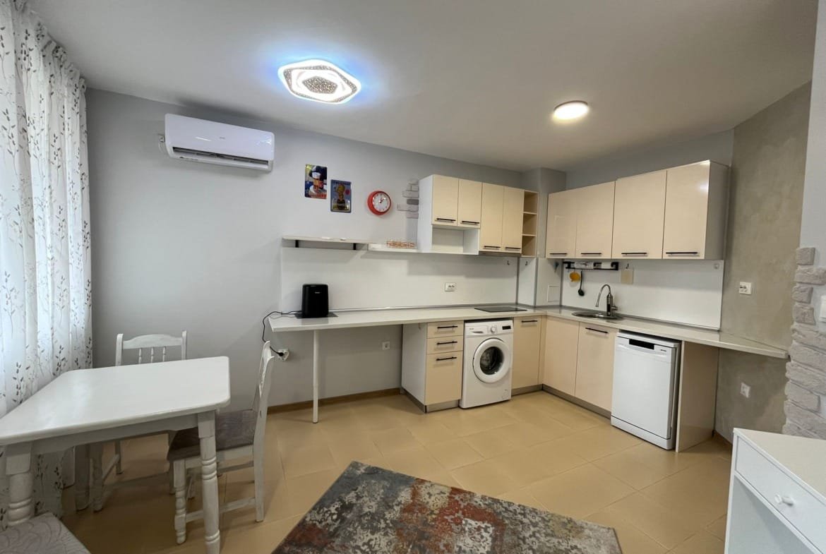 Современная кухня-студия с белыми шкафами, стиральной машиной и обеденным столом в светлых тонах, идеальна для жизни в Болгарии.