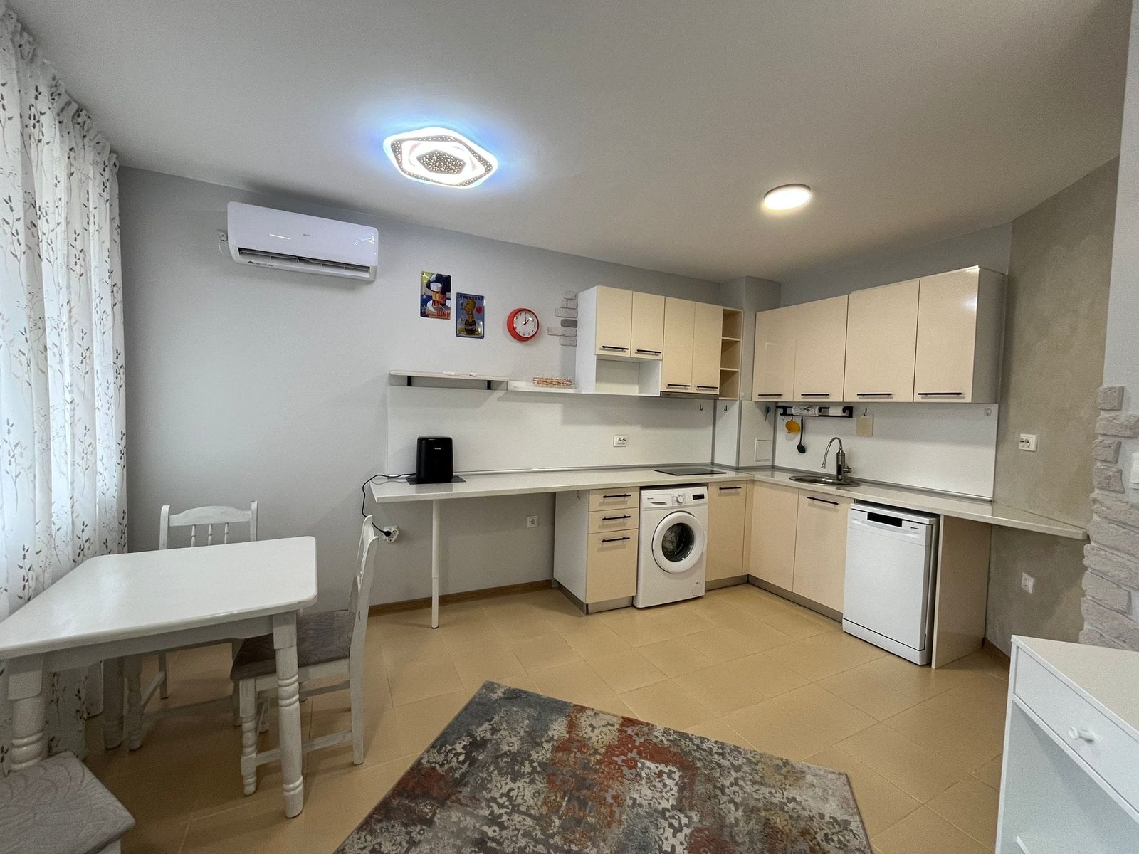 Современная кухня в светлых тонах с техникой и обеденной зоной Современная кухня-студия с белыми шкафами, стиральной машиной и обеденным столом в светлых тонах, идеальна для жизни в Болгарии.