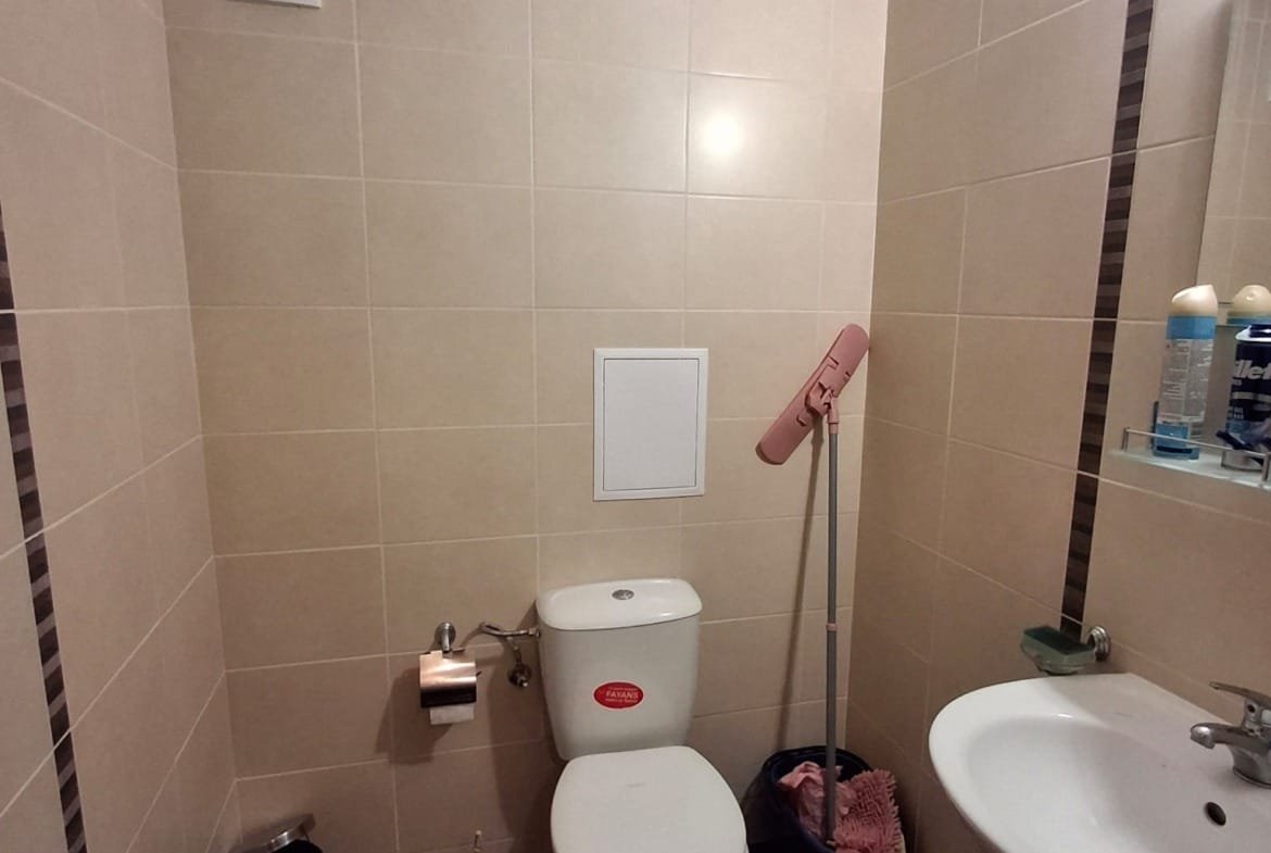 Современный туалет с плиткой, унитазом, раковиной и аксессуарами для уборки. Оборудован вентиляцией и зеркалом.