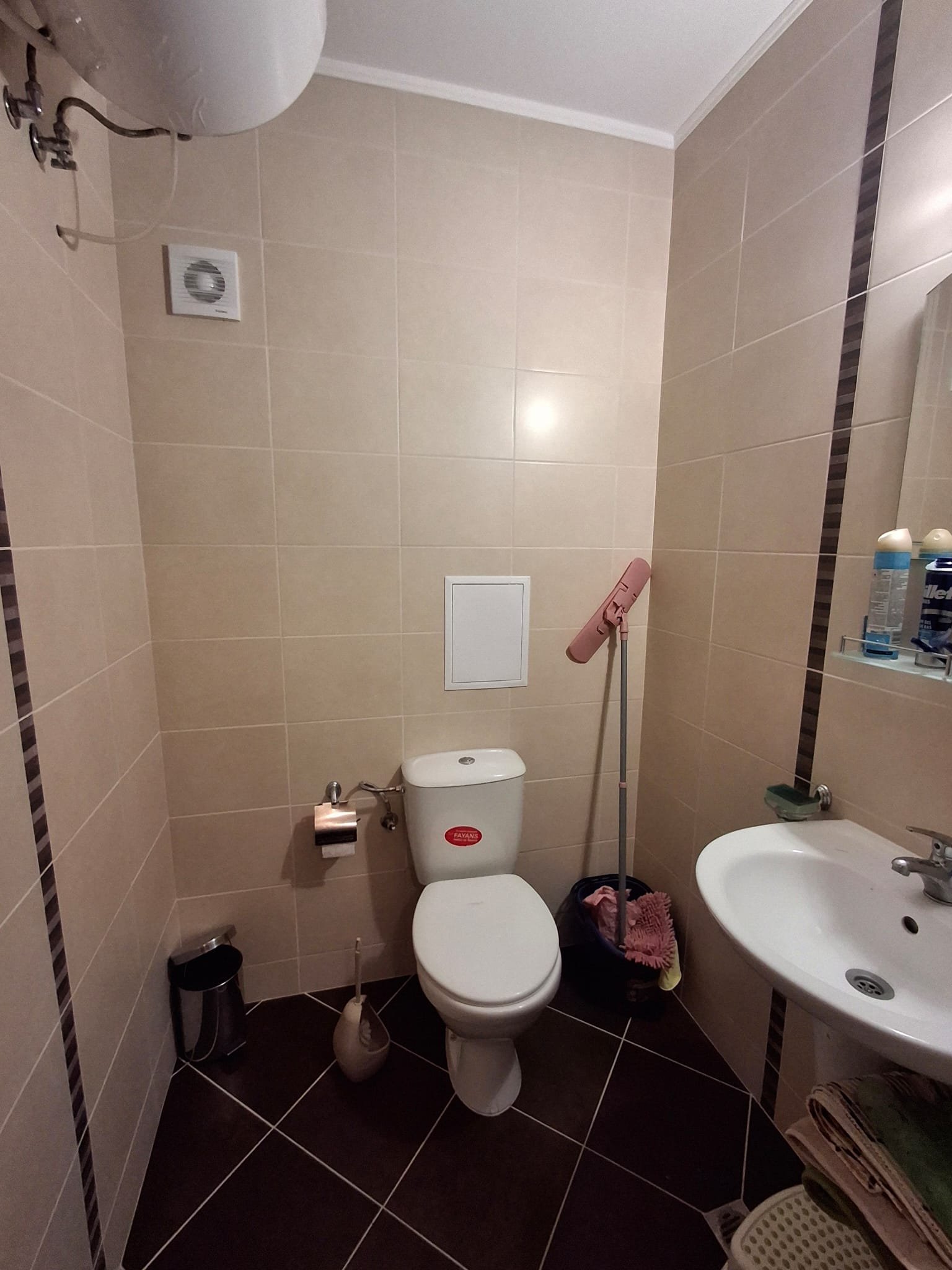 Современный туалет с плиткой, унитазом, раковиной и аксессуарами для уборки. Оборудован вентиляцией и зеркалом.