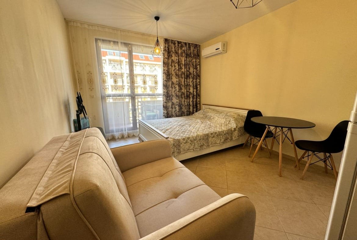 Современная квартира в Болгарии с уютной спальней, светлым интерьером и обеденной зоной у окна. Подходит для покупки или аренды.