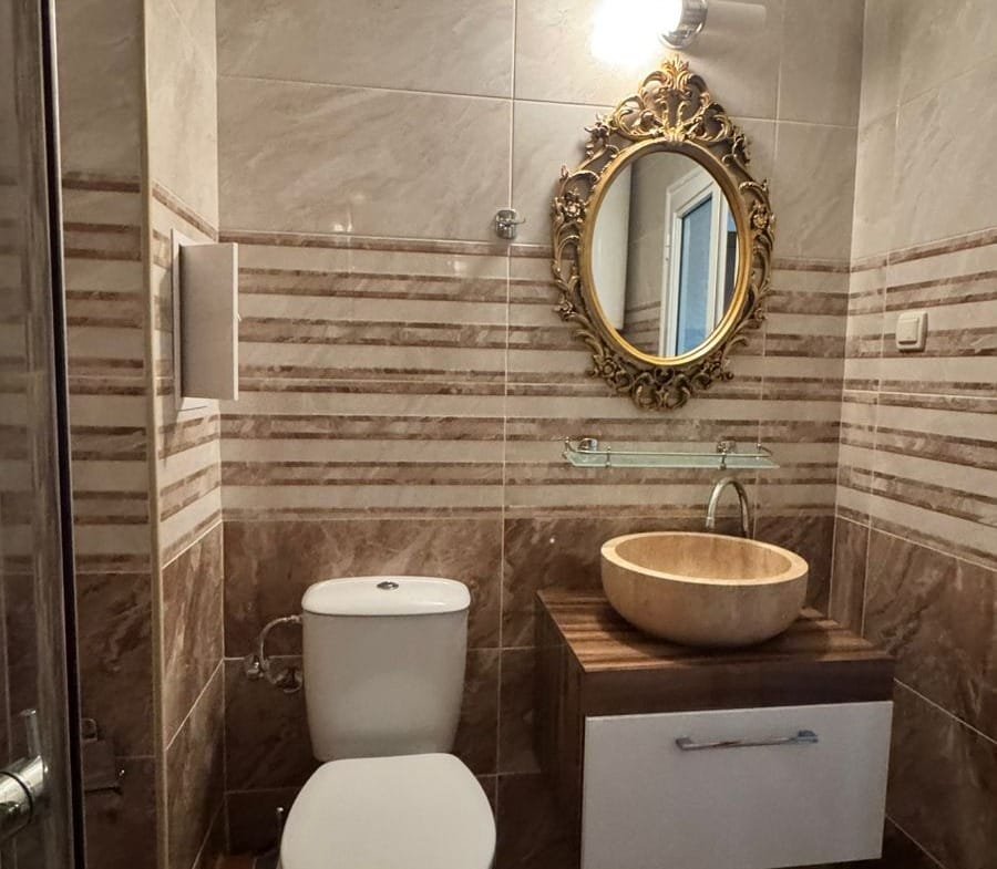 Современный санузел с стильной раковиной, унитазом и зеркалом в золотой рамке, оформленный в теплых тонах.