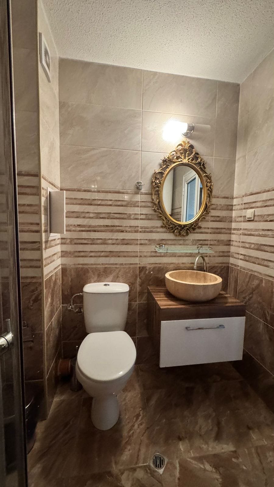 Современный санузел с стильной раковиной, унитазом и зеркалом в золотой рамке, оформленный в теплых тонах.