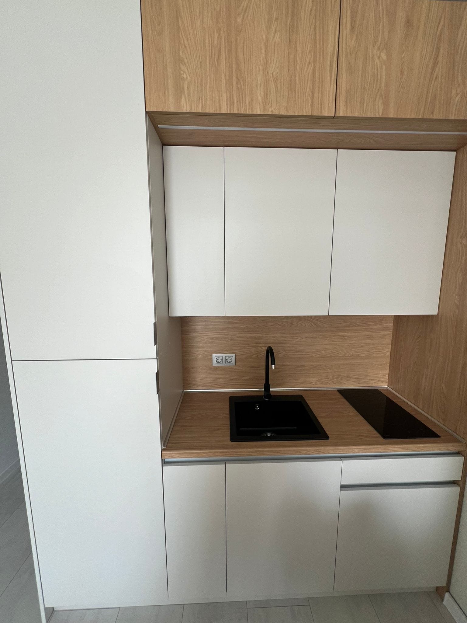 Современная кухня в стиле минимализм с белыми и деревянными фасадами, черной раковиной и варочной панелью в Болгарии.