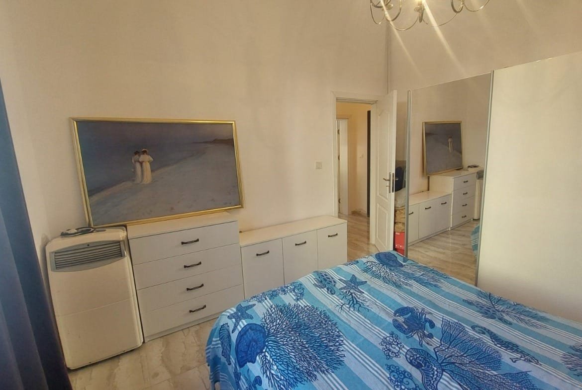 Современная спальня с зеркалом, белыми шкафами, картиной и синим текстилем, подходит для покупки недвижимости в Болгарии.