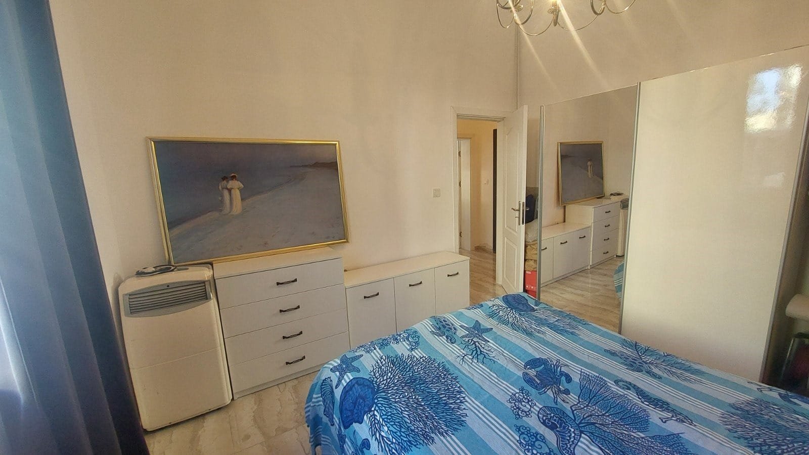 Современная спальня с зеркалом, белыми шкафами, картиной и синим текстилем, подходит для покупки недвижимости в Болгарии.