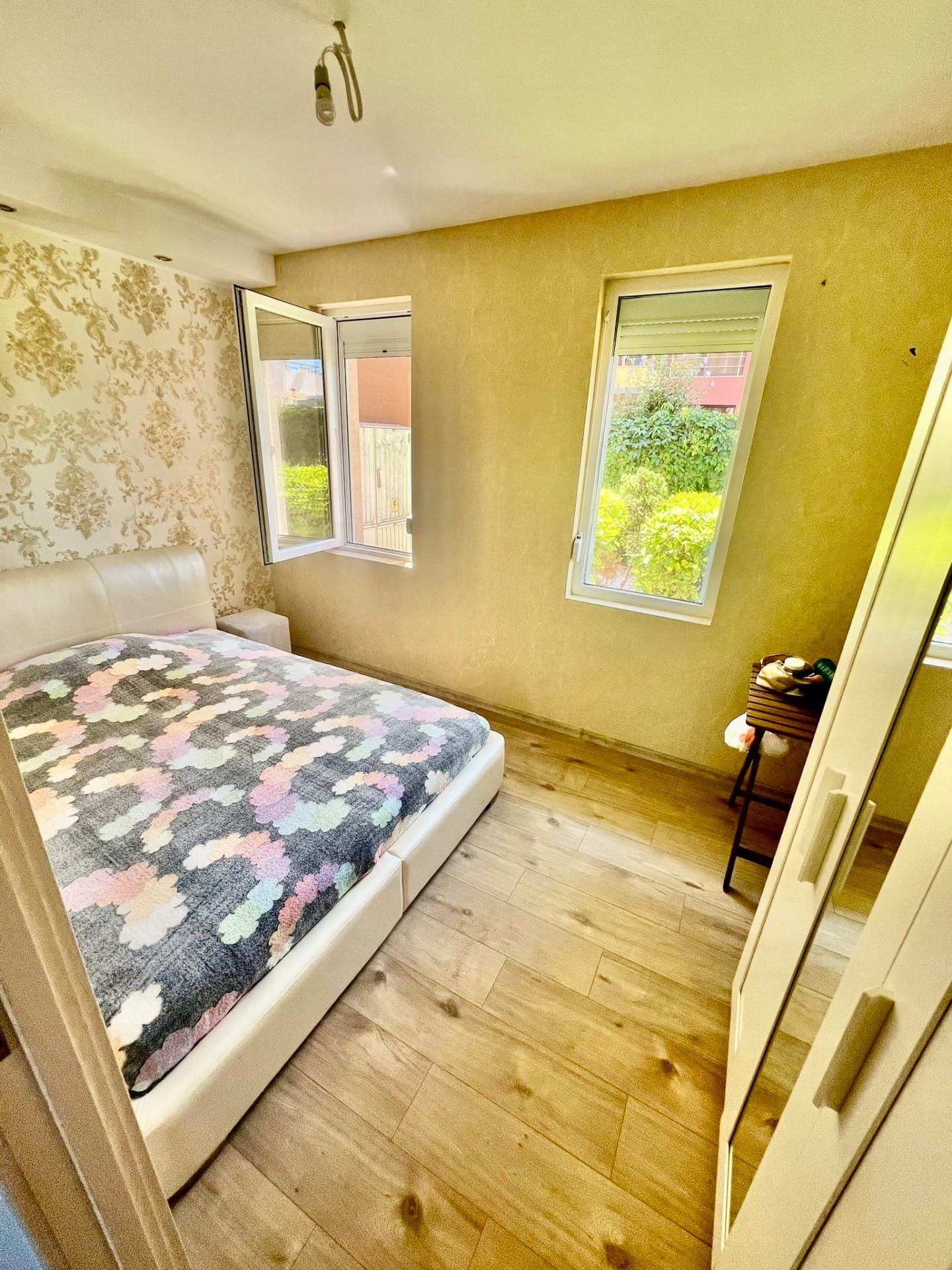 Современная спальня с белой кроватью, светлыми стенами и большими окнами, создающими уютную атмосферу для отдыха.