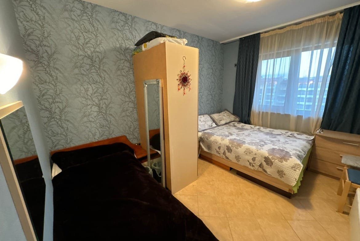 Комфортная спальня с двумя кроватями, светлыми шторами и дополнительной мебелью, идеальна для покупки недвижимости в Болгарии.