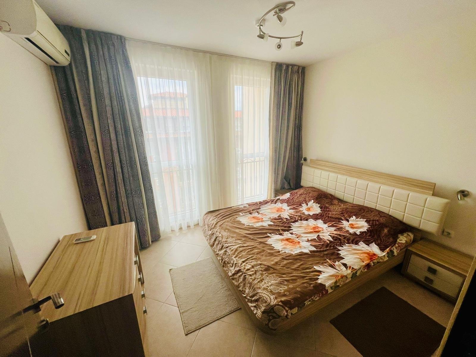 Уютная спальня с двуспальной кроватью, светлыми шторами и комодом, подходит для продажи недвижимости в Болгарии.