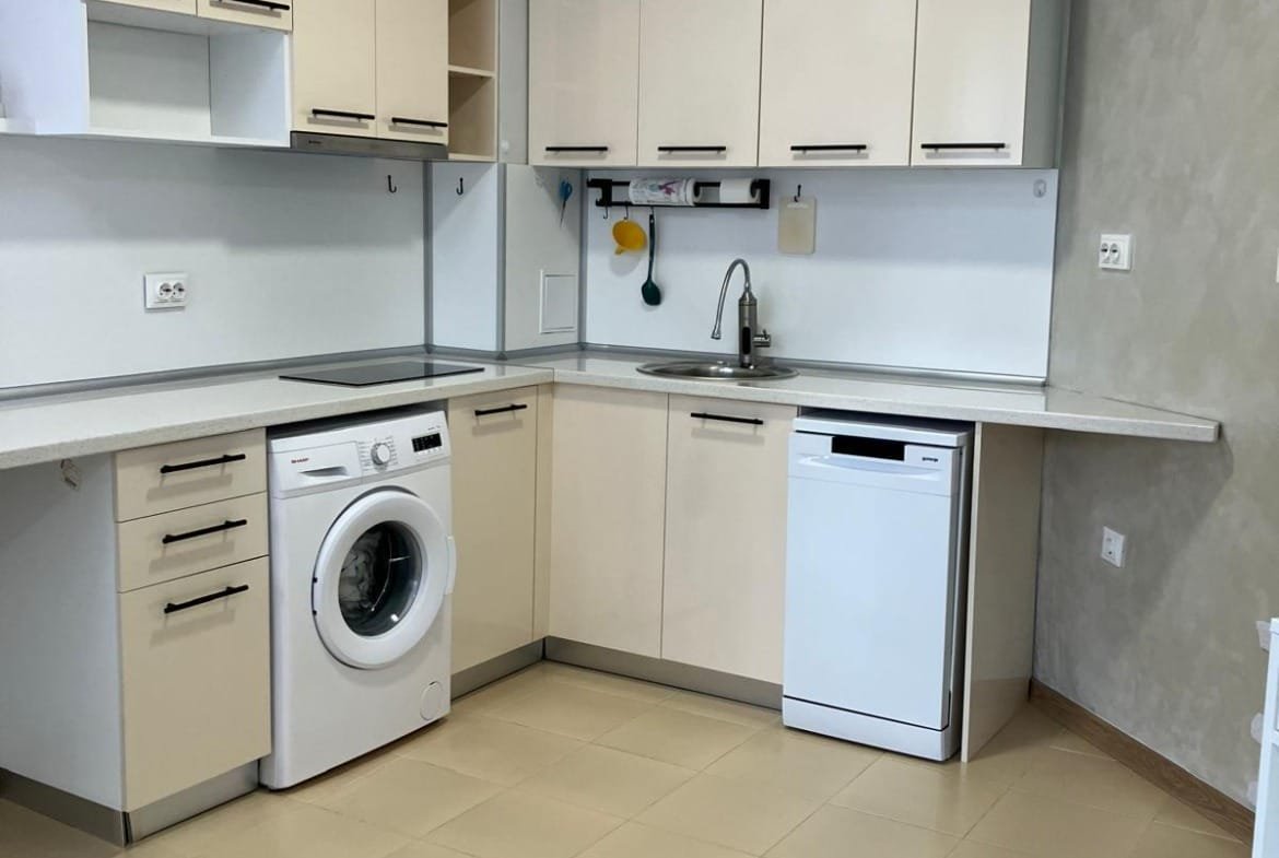 Современная кухня с бытовой техникой: стиральная машина, посудомоечная машина и светлые шкафы, подходящая для покупки недвижимости в Болгарии.