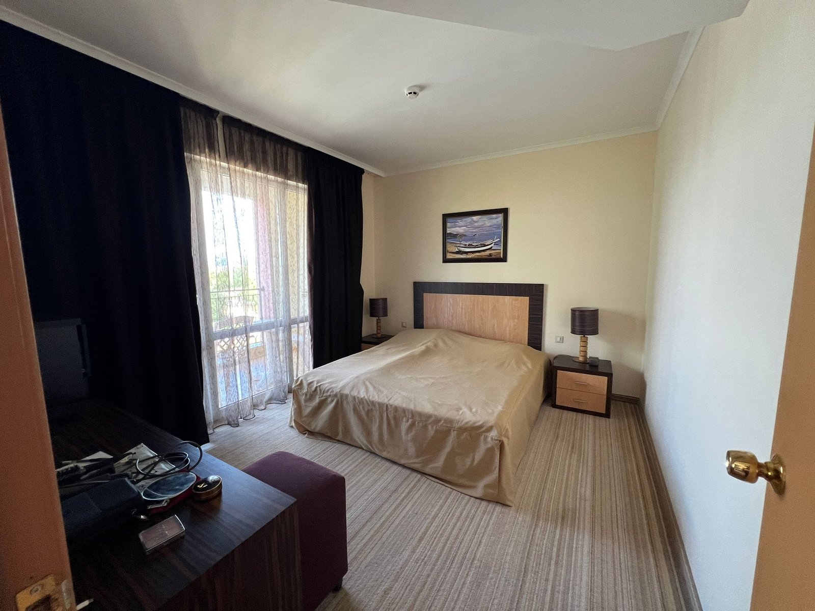 Современная спальня с двуспальной кроватью и балконом Современная спальня с двуспальной кроватью, телевизором и выходом на балкон, оформленная в светлых тонах. Идеально для покупки недвижимости в Болгарии.