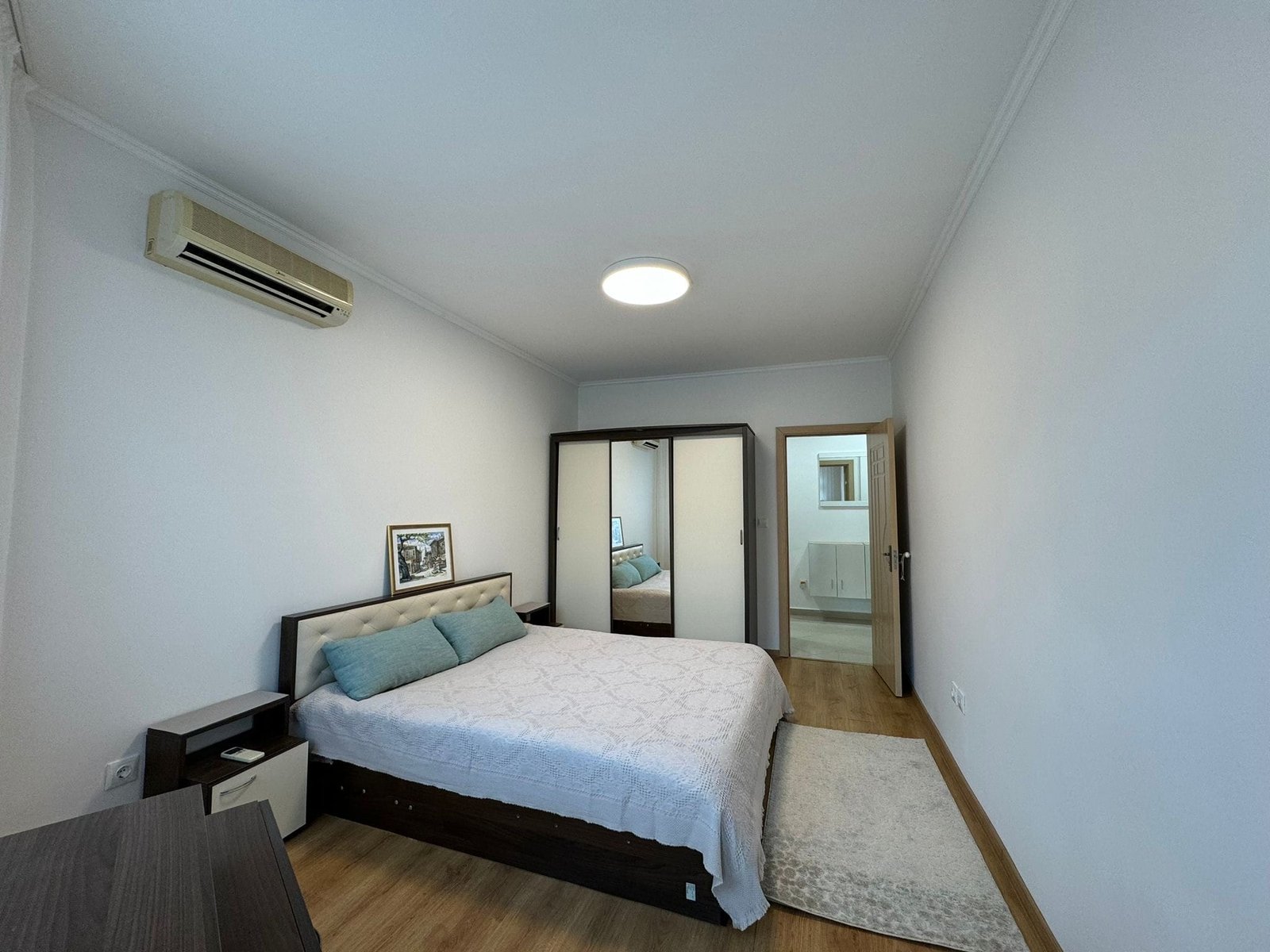 Современная спальня с двуспальной кроватью и светлыми стенами Современная спальня с двуспальной кроватью, светлыми стенами и зеркальным шкафом, идеально подходит для покупки недвижимости в Болгарии.