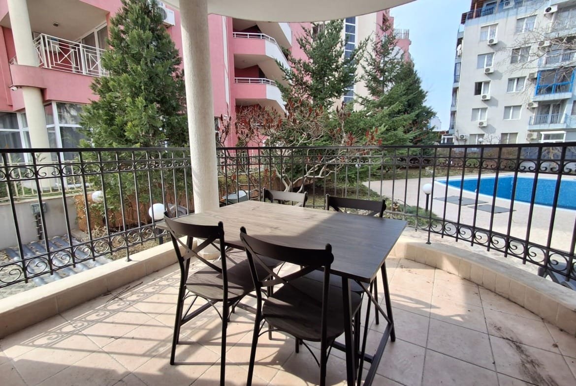 Стол и стулья на балконе с видом на бассейн и зелёные деревья, окружённые жилыми зданиями.