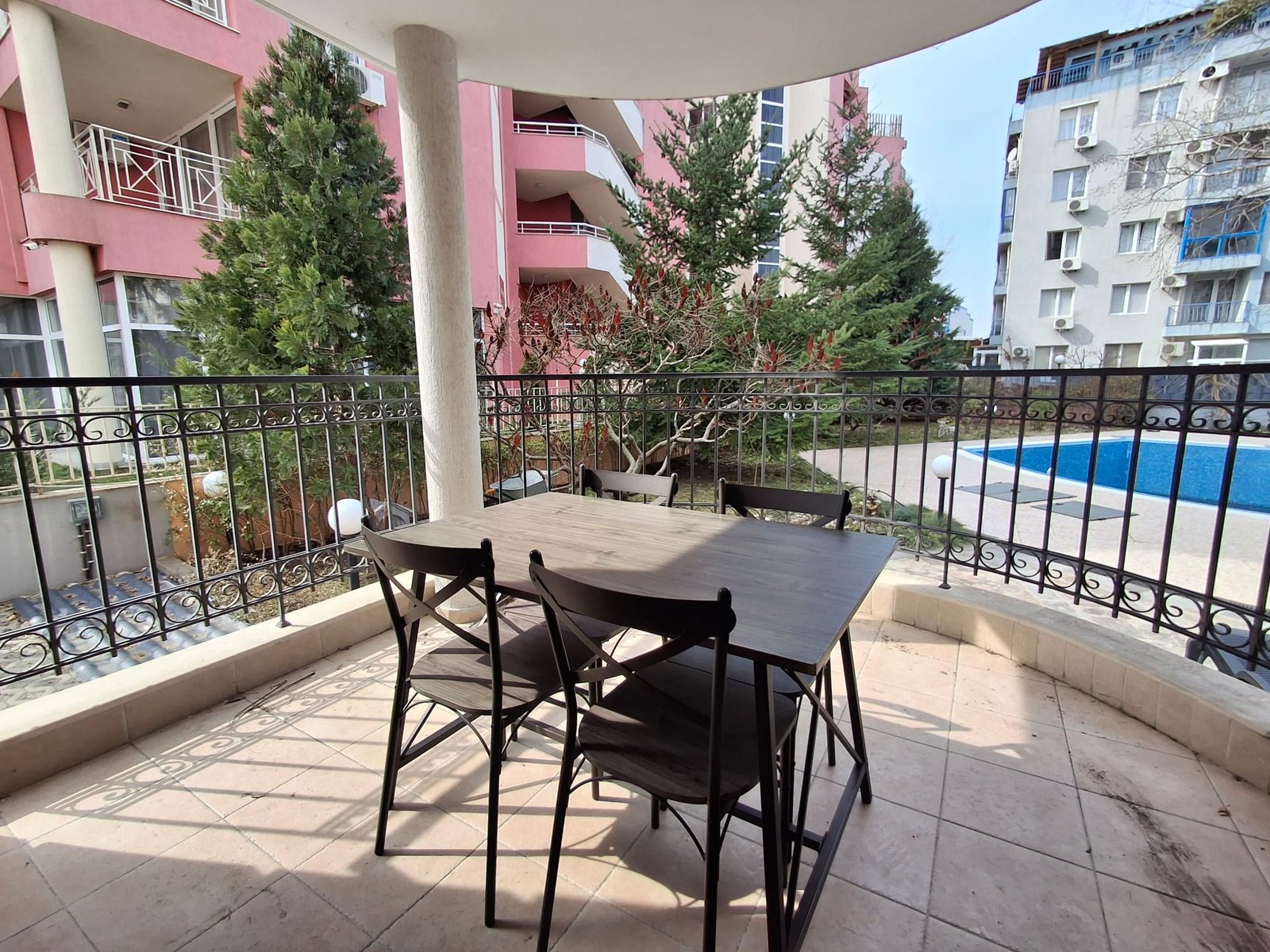 Стол и стулья на балконе с видом на бассейн и зелёные деревья, окружённые жилыми зданиями.