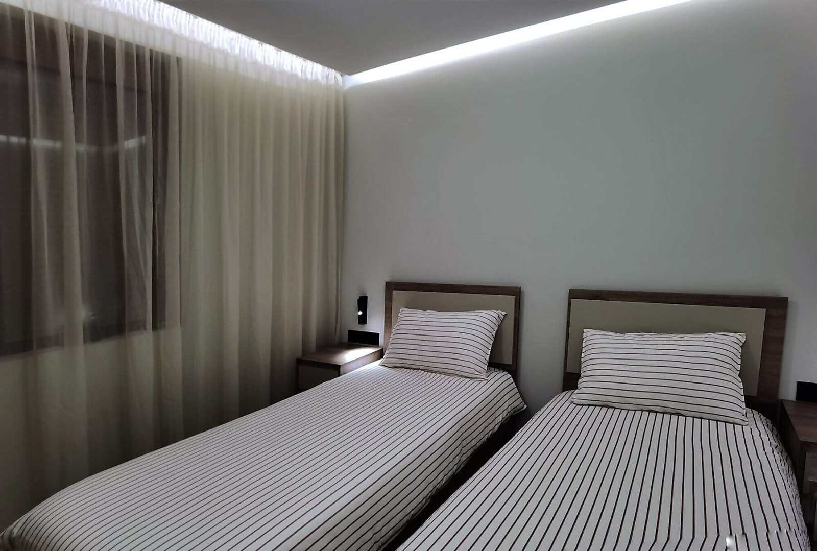 Современная спальня с двумя односпальными кроватями, светлым текстилем и мягким освещением, подходящая для жилья в Болгарии.