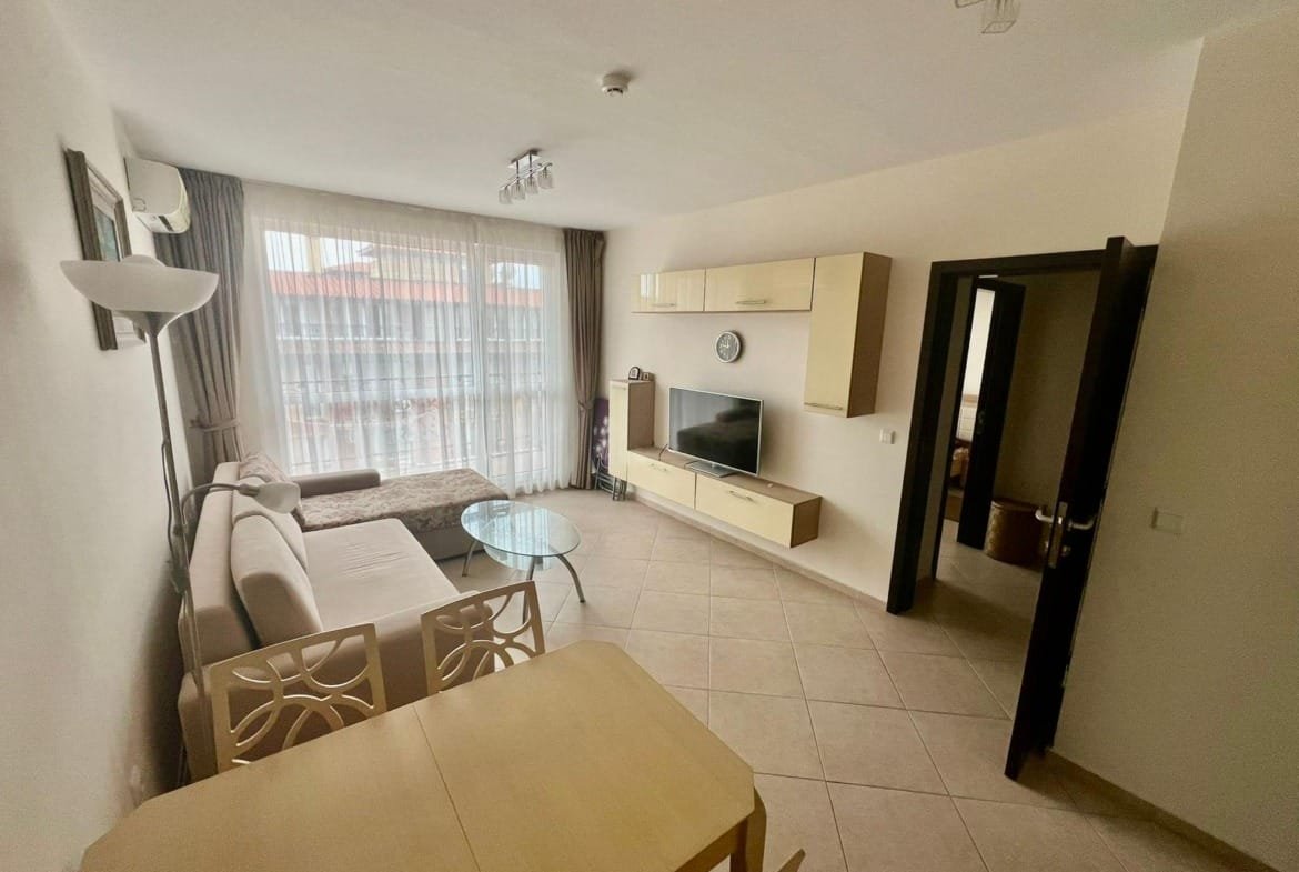 Современная квартира в Болгарии с уютной гостиной, диваном, телевизором и столом для обедов. Светлые тона, большое окно.