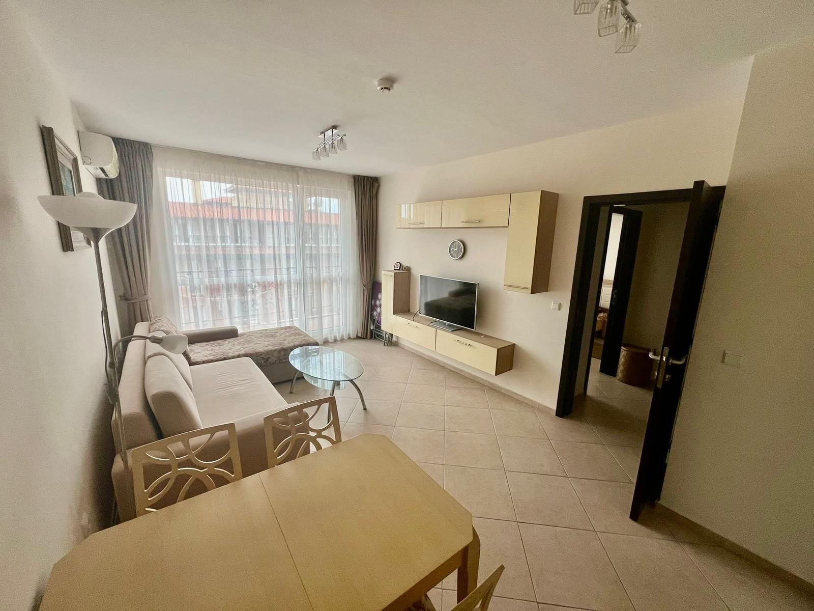 Современная квартира в Болгарии с уютной гостиной, диваном, телевизором и столом для обедов. Светлые тона, большое окно.